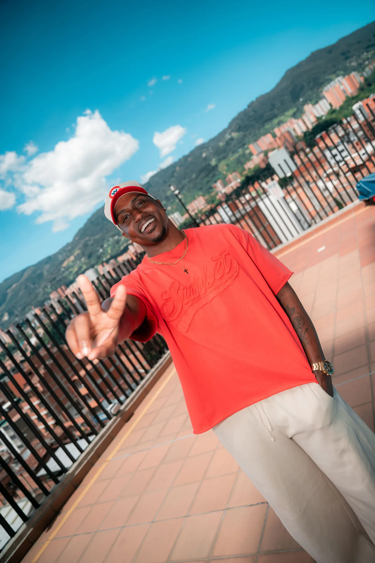 Marcus in an orange Sunset tee flashing a peace sign on a rooftop above the city